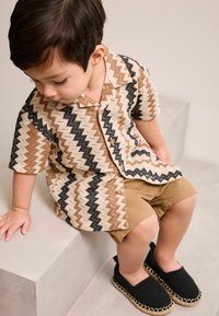 Kurzarmhemd mit Zickzackmuster in Braun, Schwarz und Creme, kombiniert mit khakifarbenen Shorts und schwarzen Espadrilles.
