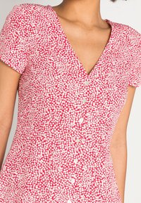 Rode jurk met een wit bloemenpatroon, V-hals, korte mouwen en knoopdetails. De stof lijkt lichtgewicht en gestructureerd.