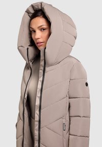 Beige polstret jakke med skrå sømme, stor hætte og lynlåslukning. Har et logostropp på siden og sorte detaljer.