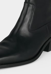 Gros plan d'une bottine noire en cuir avec un panneau latéral élastique et un talon bloc sur fond clair.