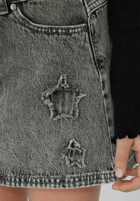 Jupe en denim mini noire et grise avec des patches en forme d'étoile usés. Elle possède un ourlet effiloché, des poches et des accents en métal.