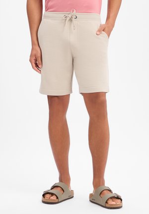 Mężczyzna ubrany w beżowe szorty ze sznurkiem, różowy top i taupe sandały na podwójnych paskach, stojący z jedną ręką w kieszeni na białym tle.