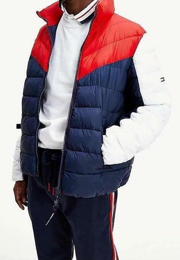 Homme portant une veste matelassée color block en rouge, bleu marine et blanc, une chemise blanche et un pantalon bleu marine avec des rayures rouges.