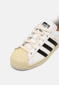 Vita lädersneakers med svarta tre ränder, beige snören och en texturerad gummitå. Guld "SUPERSTAR"-logotyp på sidan.