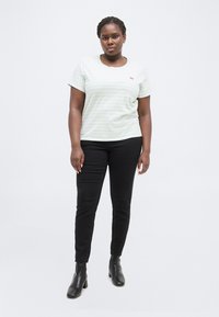 Γυναίκα που στέκεται με το πρόσωπο προς τα εμπρός, φορώντας ελαφρώς ριγωτό κοντομάνικο πουκάμισο, μαύρο παντελόνι και μαύρα ανδρικά μποτάκια με τακούνι, σε λευκό φόντο.