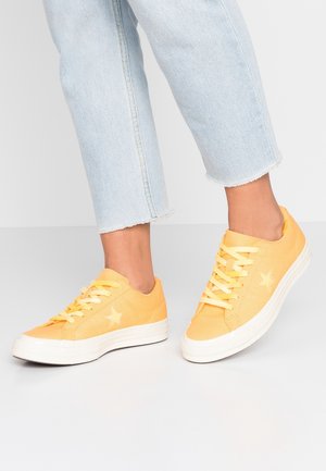 Personne portant un jean court bleu clair et des baskets jaunes à lacets avec des semelles blanches et des logos en forme d'étoile.