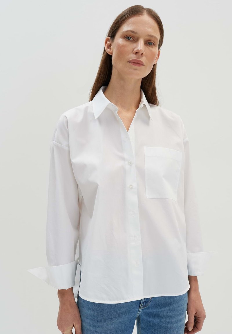 Camicia bianca in cotone con taschino sul petto, maniche ripiegate e vestibilità comoda. Texture morbida, design classico del colletto e orlo curvo.