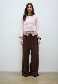 Mlada ženska stoji ob običajni steni, oblečena v roza dolg rokav čez črtasto majico in široke rjave hlače ter temne čevlje.