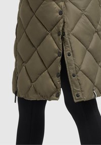 Olive green quilted jacket with black snap buttons, side zipper, worn over black ribbed leggings showing mid-thigh area.