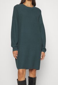 Robe tricotée teal avec une texture côtelée, manches longues, col rond et coupe décontractée. Associée à des bottes noires montantes.