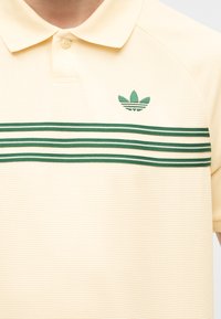 Μπεζ πόλο πουκάμισο με πράσινο λογότυπο Adidas trefoil στην αριστερή πλευρά του στήθους και τρεις οριζόντιες πράσινες ρίγες στο στήθος.