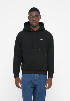 Black hoodie made of cotton with a front pocket, drawstring hood, and white "OBEY" logo on the chest. Paired with blue jeans.