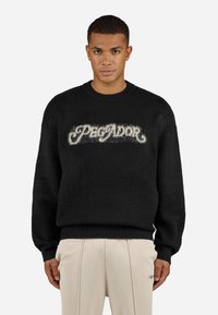 Black sweater made of fuzzy material, featuring a prominent cream logo "PREDATOR" in a decorative font on the front and ribbed cuffs.