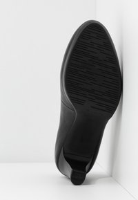Chaussure de ville noire inclinée vers le haut, montrant une semelle antidérapante à motif et un petit talon sur fond blanc d’angle.