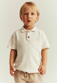 Helles beige Polohemd mit kurzen Ärmeln, strukturiertem Muster, drei Knöpfen und einer Brusttasche, kombiniert mit hell gestreiften Hosen.