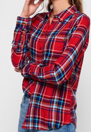 Žena v červené, modré a bílé kostkované košili na knoflíky s dlouhými rukávy a modrými džínami, pózující s jednou rukou u obličeje.
