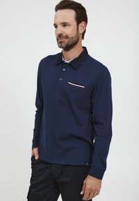 Dlhý rukáv, námornícky modrý polo tričko s kontrastným čiernym golierom, tromi gombíkmi a náprsným vreckom so červenými a bielymi prúžkami.