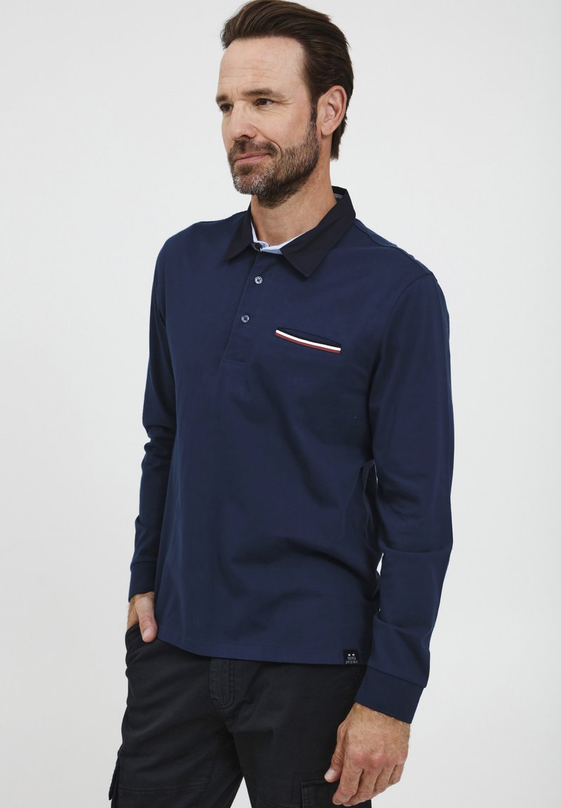 Dlhý rukáv, námornícky modrý polo tričko s kontrastným čiernym golierom, tromi gombíkmi a náprsným vreckom so červenými a bielymi prúžkami.