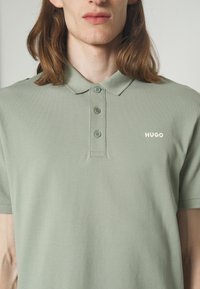 Gros plan d'une personne portant un polo vert clair avec trois boutons et le mot HUGO imprimé sur la poitrine.