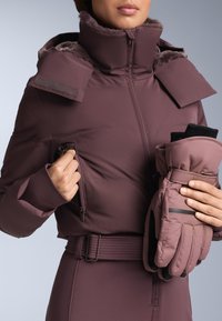 Bordeauxrode geïsoleerde jas met een hoge kraag, ritssluitingen aan de zakken en een tailleband met riem, gecombineerd met bijpassende gewatteerde handschoenen met zwarte accenten.