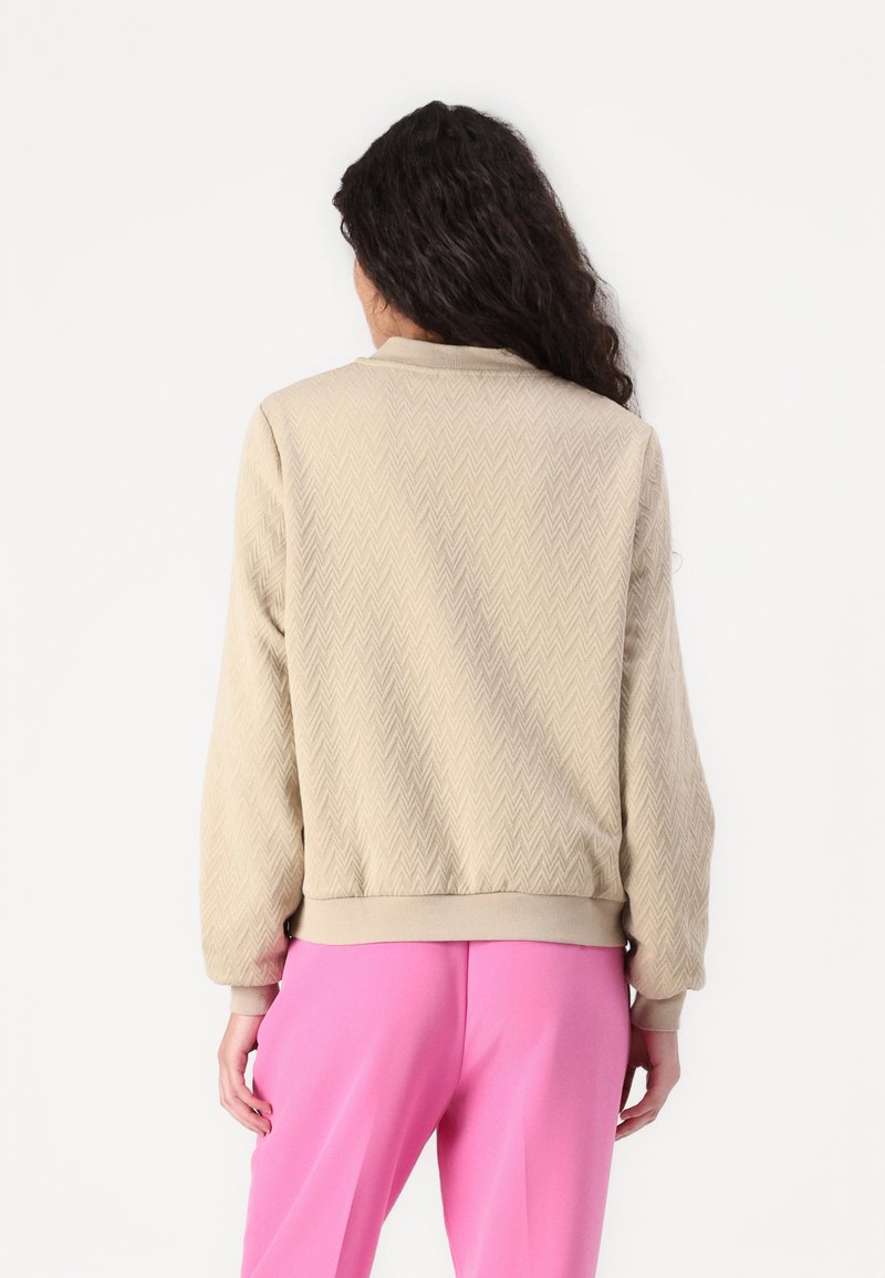 Giacca bomber beige con una trama a spina di pesce testurizzata, polsini e orlo a costine. Abbinata a pantaloni rosa brillante, che mostrano una vestibilità rilassata.