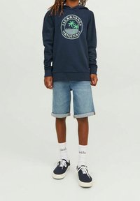 Marinblå hoodie med cirkulärt logotyp, jeanstightshorts med uppvikta ändar, vita strumpor och marinblå sneakers med vita sulor. Avslappnad outfitdesign.