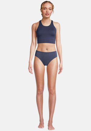 Junge Frau steht barfuß, trägt ein dunkelblaues bauchfreies Oberteil und passende hochgeschnittene Unterwäsche, vor einem schlichten weißen Hintergrund.