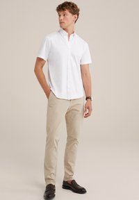 Camisa blanca de manga corta con botones, combinada con pantalones beiges y mocasines oscuros, con un cuello estructurado y una textura de tela suave.