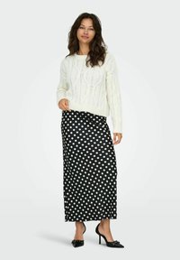 Maglione bianco a trecce abbinato a una gonna maxi nera a pois, caratterizzata da una texture liscia, vita alta e tacchi neri con dettagli a fiocco.