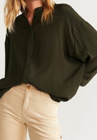 Blusa verde oscuro, holgada, con cuello mao, botonera en la parte delantera y mangas anchas, combinada con pantalones de pana color beige claro.