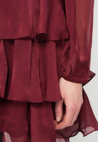 Δεξί χέρι resting δίπλα σε πολύχρωμο sheer μπορντό ύφασμα με ρυτίδες και μανσέτα από ελαστικό στον ανοιχτό φόντο.