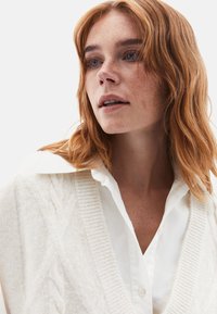 Cardigan en tricot torsadé crème superposé à une chemise blanche à col. Taches de rousseur sur le visage, cheveux ondulés rouges et arrière-plan neutre.