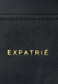 Svart lädermaterial med en texturerad yta, med varumärket "EXPATRIÉ" präglat i guld. Stygn längs kanten.