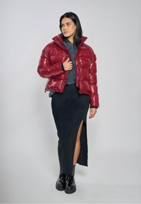 Röd glansig vadderad jacka med hög krage och synliga sömmar, kombinerad med en svart ribbad kjol med sidospjäll och svarta kraftiga stövlar.