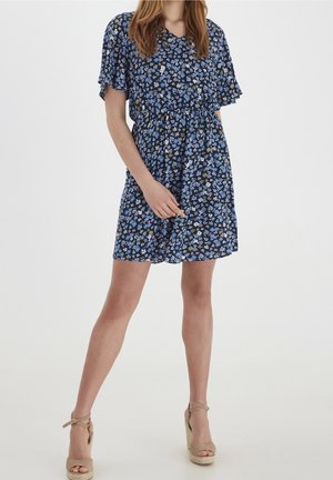 Žena v krátkých černých šatech s modro-bílým květinovým potiskem a béžovými vysokými sandály na platformě, stojící s nohama od sebe.