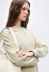 Νεαρή γυναίκα με μακριά καστανά μαλλιά, φορώντας μπεζ φούτερ και κρατώντας μια τσάντα ώμου στον ίδιο τόνο, με σταυρωμένα χέρια, κοιτώντας προς τα πλάγια.