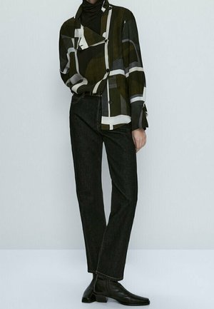 Personne portant une veste à motif géométrique, un col roulé noir, un jean droit foncé et des bottines noires sur un fond uni.