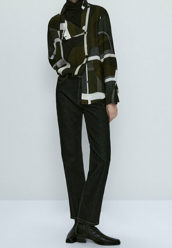 Personne portant une veste à motif géométrique, un col roulé noir, un jean droit foncé et des bottines noires sur un fond uni.