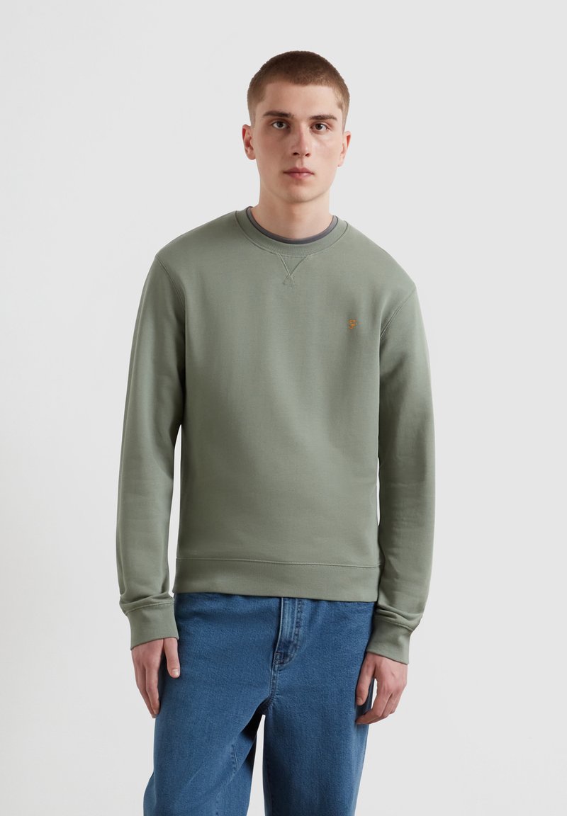 Olijfgroene sweatshirt met een ronde halslijn en geribbelde mouwen, met een klein geborduurd logo op de borst. Draagt bij blauwe spijkerbroek.