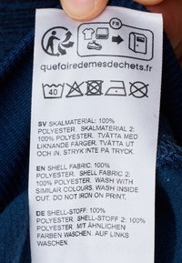 Etichetta di cura su tessuto con istruzioni per la cura in più lingue. Tessuto bianco con testo nero, inclusi simboli di lavaggio e link al sito web.
