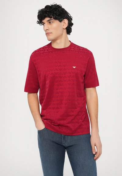Rdeča majica s kratkimi rokavi z geometričnim vzorcem in okroglim izrezom. Majhen logotip na prsih, kombinirano s temnimi kavbojkami.