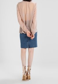 Blusa a maniche lunghe trasparente di colore rosa chiaro con polsini arricciati, abbinata a una gonna di jeans blu. Il modello indossa stivali lucidi con tacco alto e fino al ginocchio.