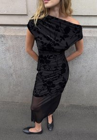 Black dress with off-shoulder design, textured floral pattern, and fitted silhouette. Sheer fabric accents at the hem and flat black shoes.