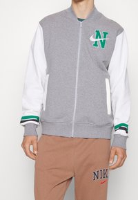 Giacca grigia con zip e maniche bianche, polsini a strisce verdi e nere, con un logo 'N' verde e il logo Nike bianco sul petto.