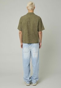 Chemise kaki à manches courtes avec un motif de feuilles texturé, associée à un jean baggy bleu clair et des baskets beiges, vue de l'arrière.