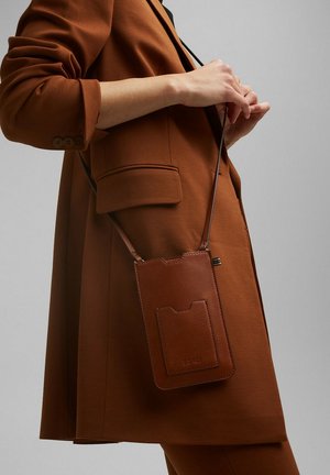 Étui de téléphone en cuir marron avec une finition texturée, doté d'une poche avant et d'une sangle réglable, tenu contre une tenue de couleur similaire.