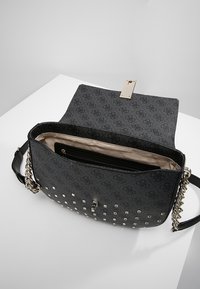 Sac à main noir à motif avec une finition texturée, orné de clous argentés, de ferrures dorées et d'une bandoulière chaîne. L'intérieur comprend une poche zippée.