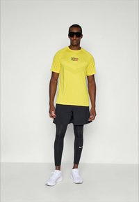Gul atletisk t-skjorte med "BERLIN RUN" trykk, i kombinasjon med svarte shorts og leggings. Hvite sneakers fullfører det sporty utseendet.