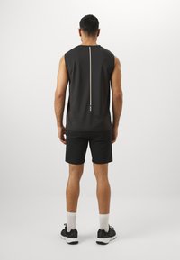 Top negru fără mâneci, cu o cusătură centrală de culoare deschisă și detalii cu logo, asortat cu pantaloni scurți negri și pantofi sport negri.