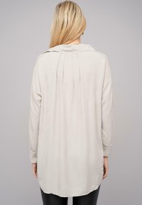 Blusa beige a maniche lunghe con vestibilità rilassata, caratterizzata da uno schienale plissettato, colletto e tessuto morbido e testurizzato. Orlo curvo e maniche con bottoni.